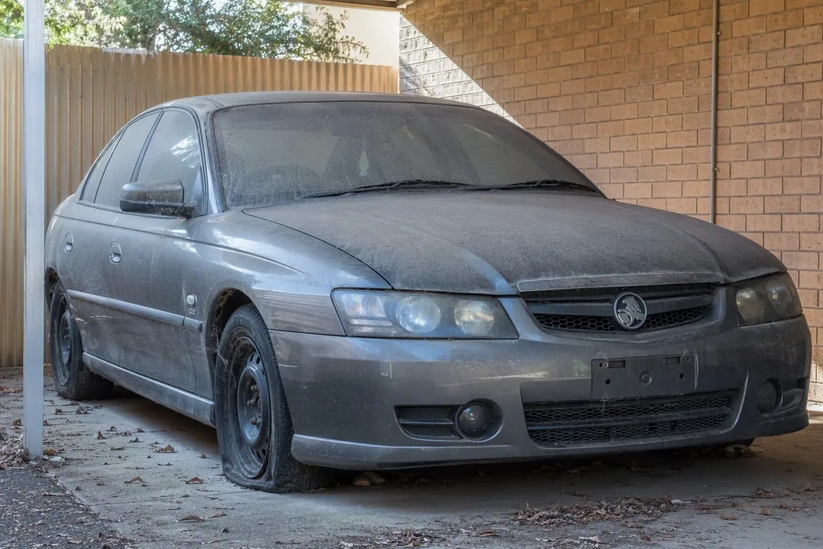 Dusty long-parked sedan with no registration plates sitting near a suburban shed